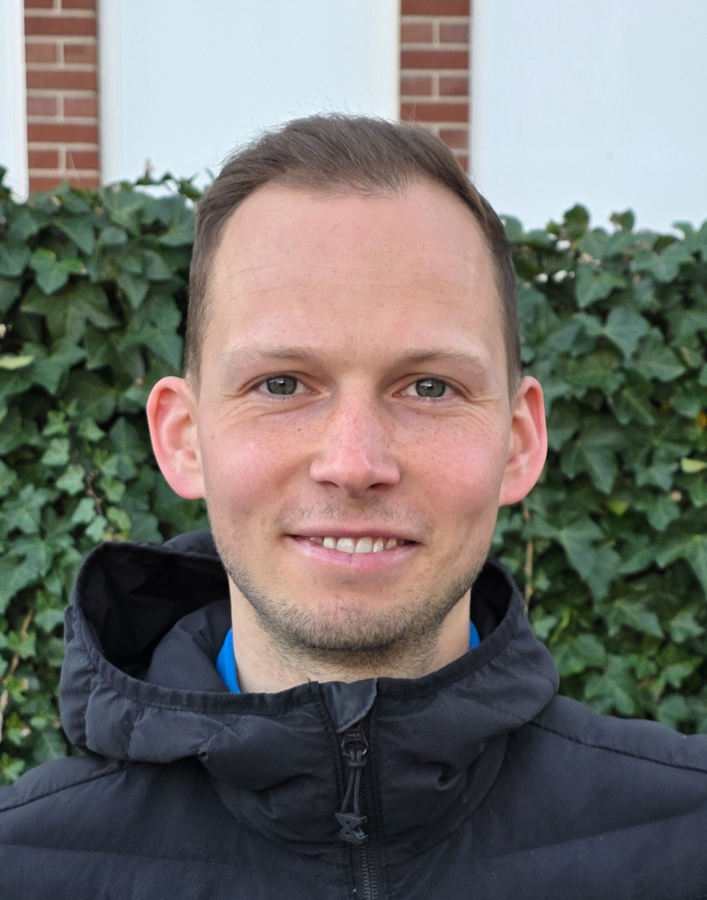 Eine lächelnde Person im schwarzen Mantel vor Ivy-Wand, kurzer dunkler Haaransatz.