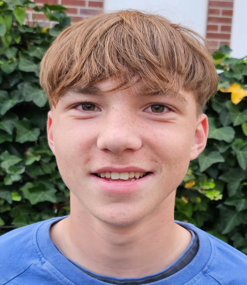 Laechelnder junger Mann mit blonden Haaren und blauem Shirt vor gruenem Efeu