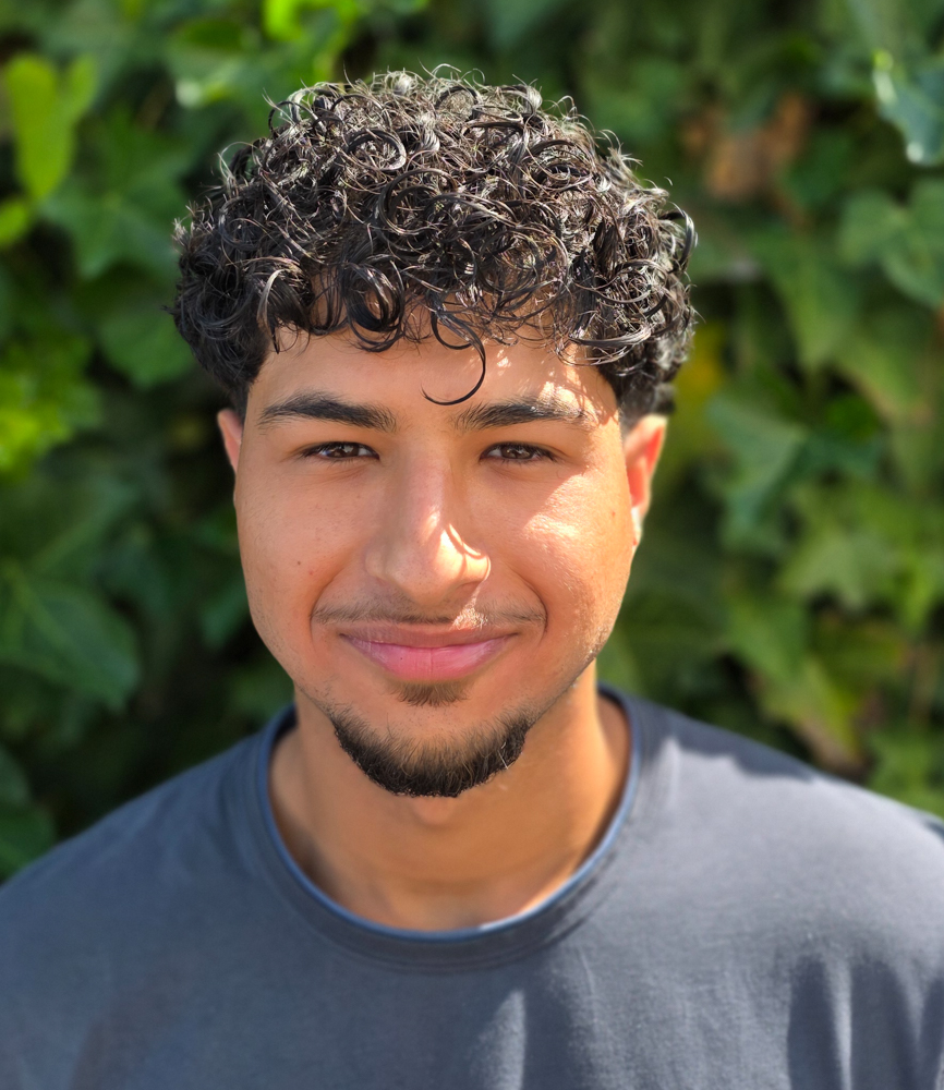 Junger Mann mit dunklen Locken laechelt leicht im grauen Shirt vor Gruen